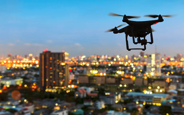 quadcopter drone in silhouette flying high above a brightly lit cityscape at twilight